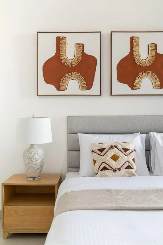 Una cama bien hecha con cabecero gris está bajo dos cuadros enmarcados.