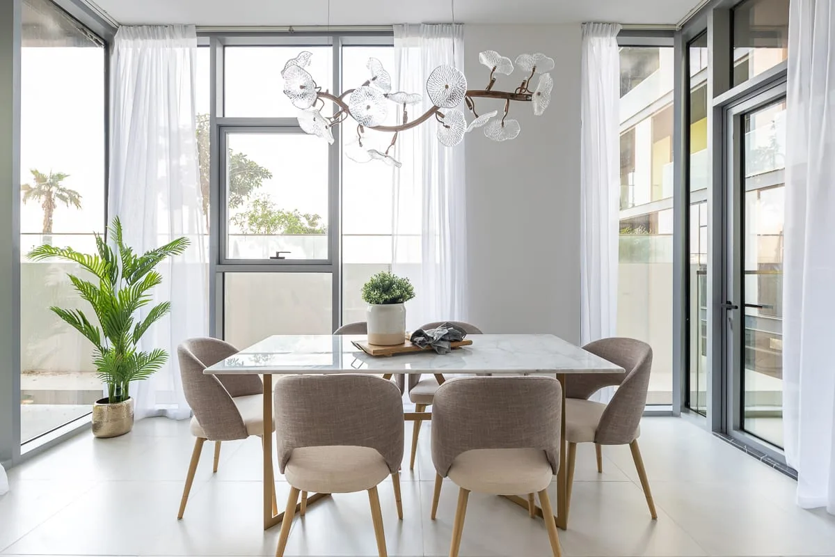 Un comedor luminoso con mesa, asientos tapizados y grandes ventanales.