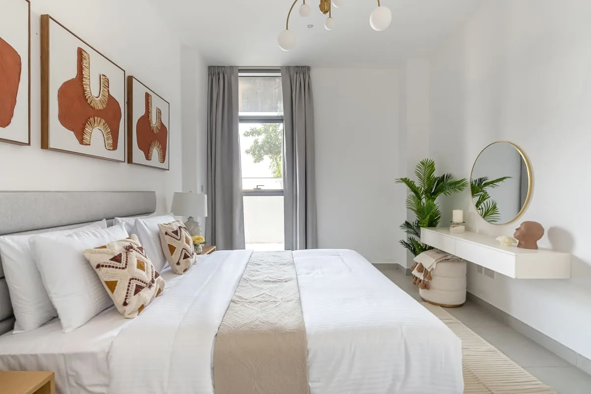Un dormitorio luminoso con ropa de cama blanca, cuadros enmarcados y una ventana con cortinas grises.