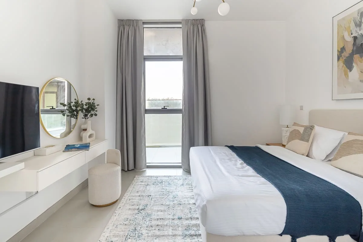 Un dormitorio luminoso con una cama, cortinas grises y un televisor montado en la pared.