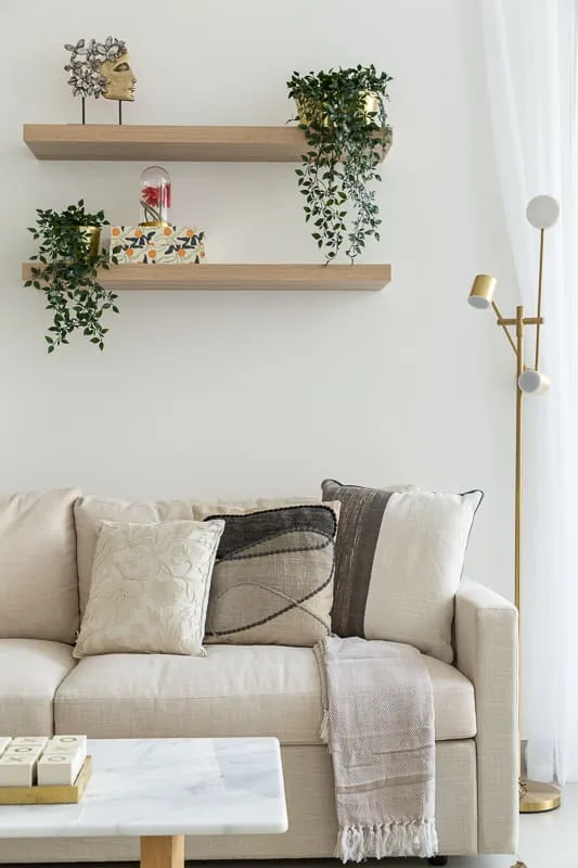 Un sofá claro está bajo estantes de madera con plantas en una sala luminosa.