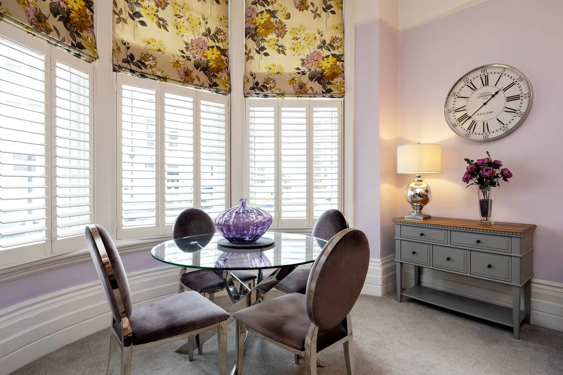 Comedor con mesa de cristal, asientos tapizados y ventanas luminosas con persianas.