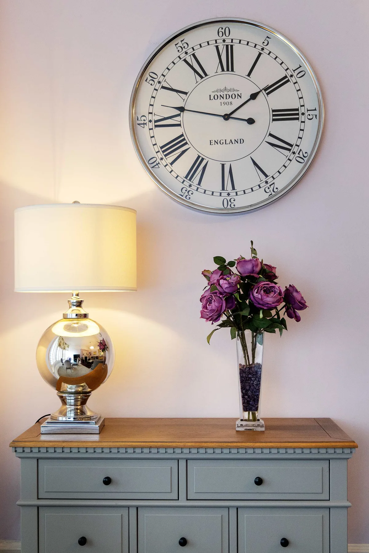 Un reloj decorativo cuelga sobre una consola con una lámpara y flores.