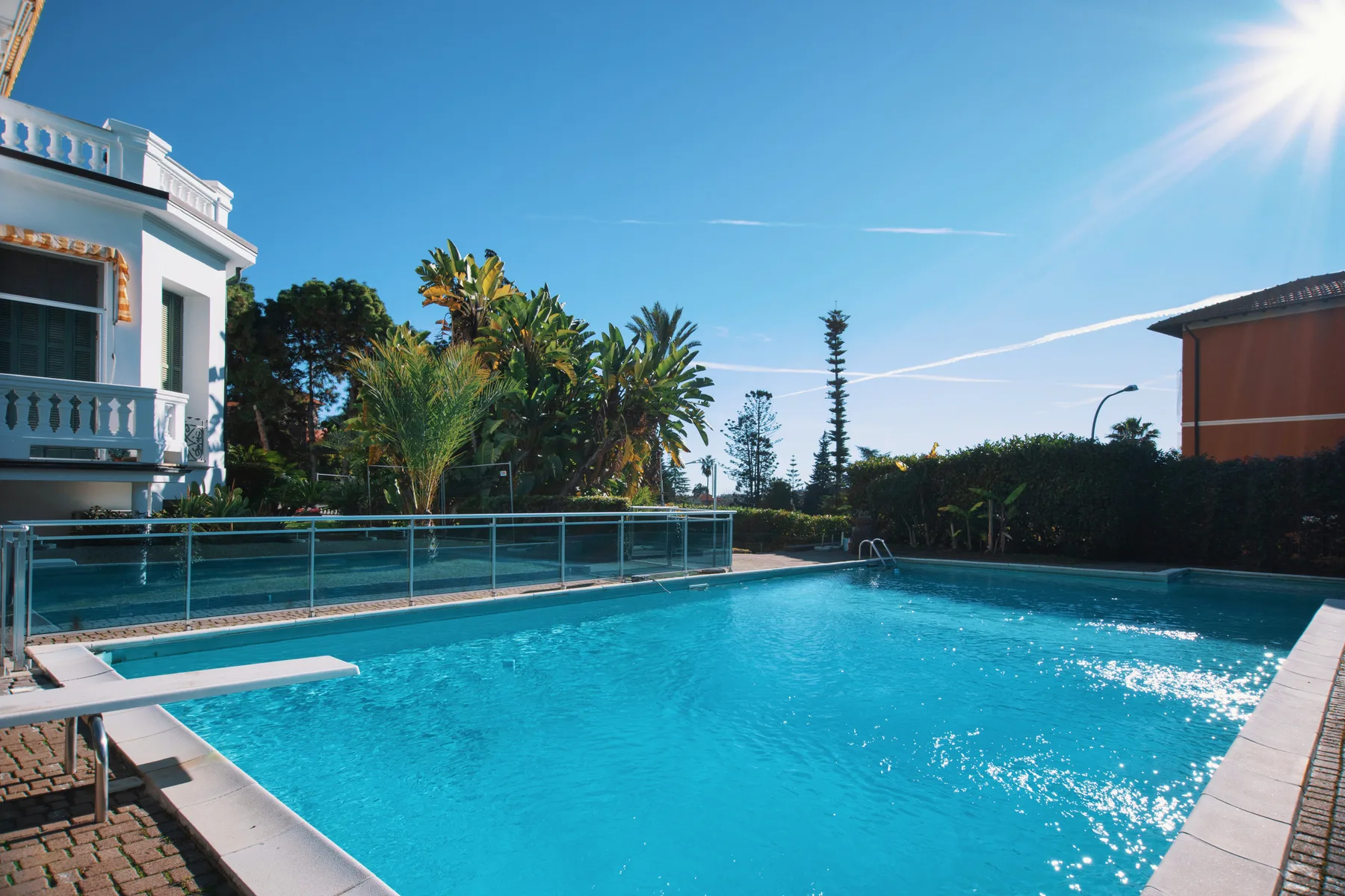 Una piscina exterior iluminada por el sol está rodeada de vegetación y edificios cercanos.