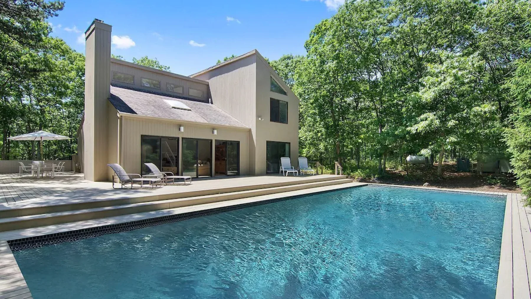 A modern house overlooks a swimming pool, with leafy trees surrounding the yard.