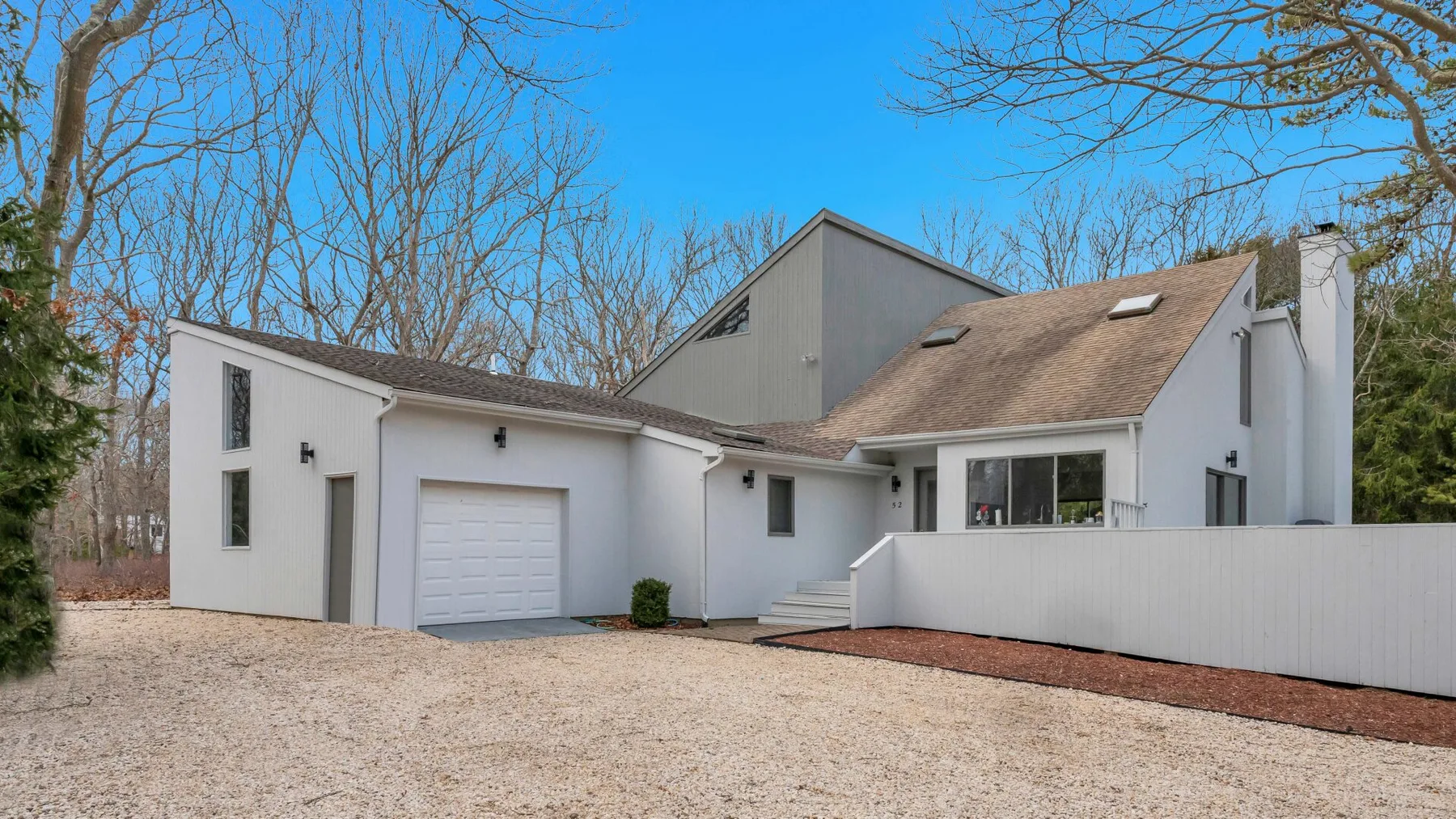 A white modern house stands beside a broad gravel driveway and leafless trees.