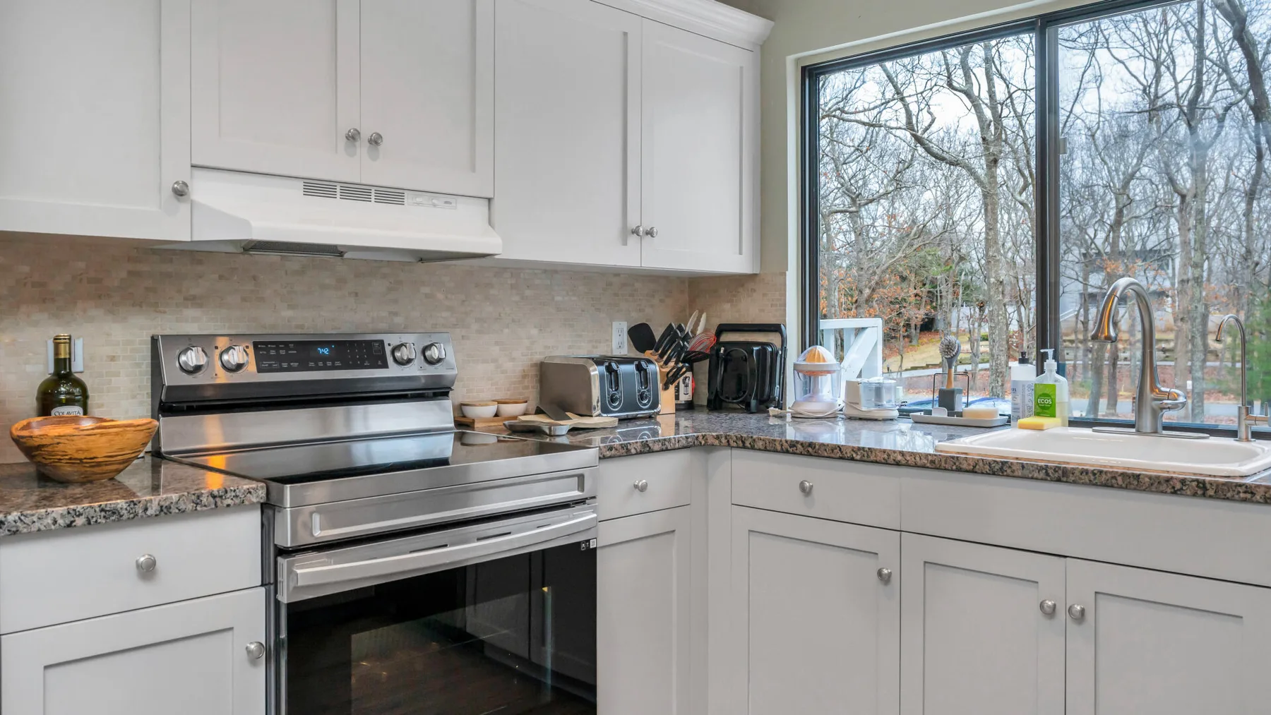 Una cocina luminosa con gabinetes blancos, una estufa y una gran ventana con vista a los árboles.