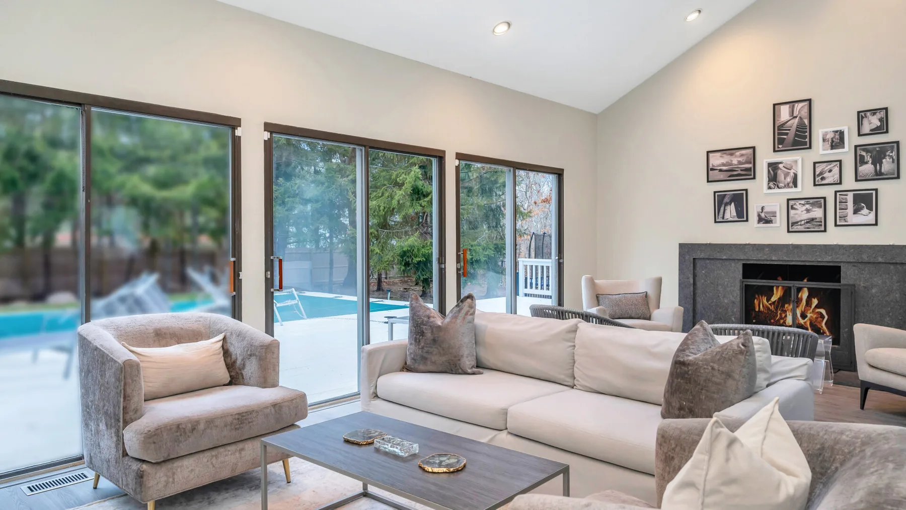 Una sala luminosa tiene asientos claros, una mesa de centro y grandes ventanas con vista a una piscina.