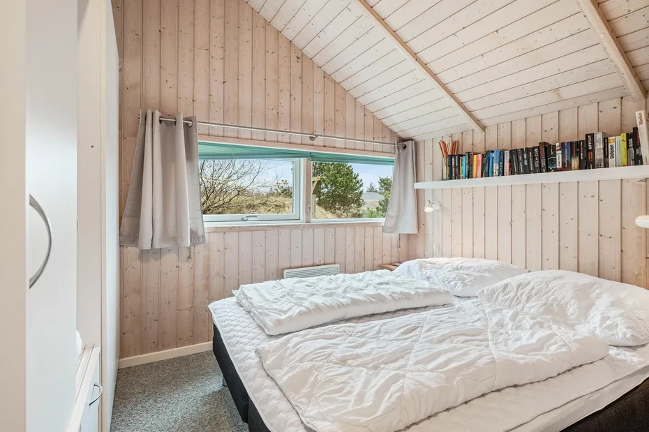 A bedroom with a bed, a window, and books on a wall shelf.