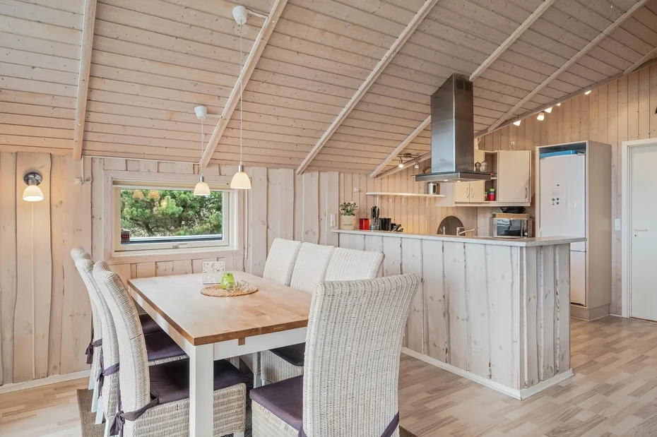 A bright dining area opens onto a kitchen, with a wooden table and woven seating.