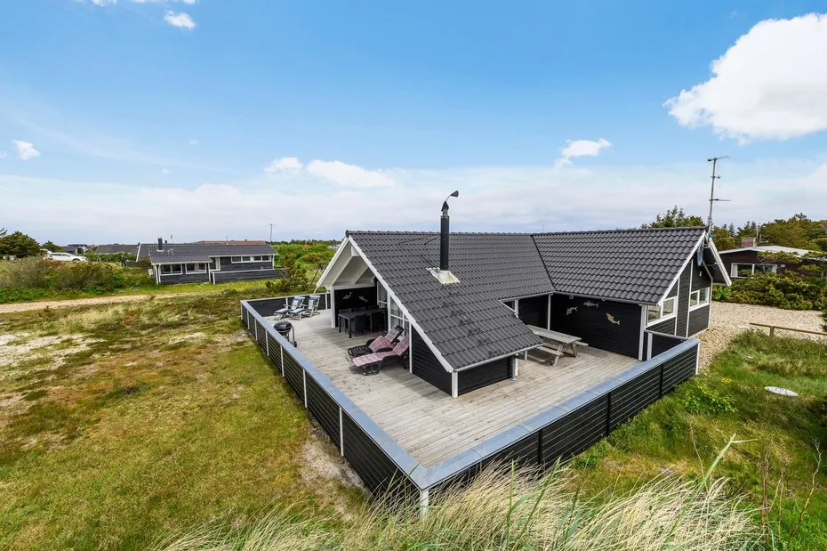 A dark-roofed house sits on a broad wooden deck surrounded by open grass.