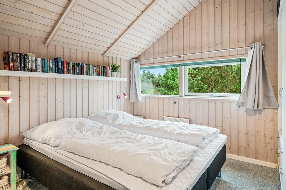 A bedroom with a double bed beneath a sloped wooden ceiling and a wide window.