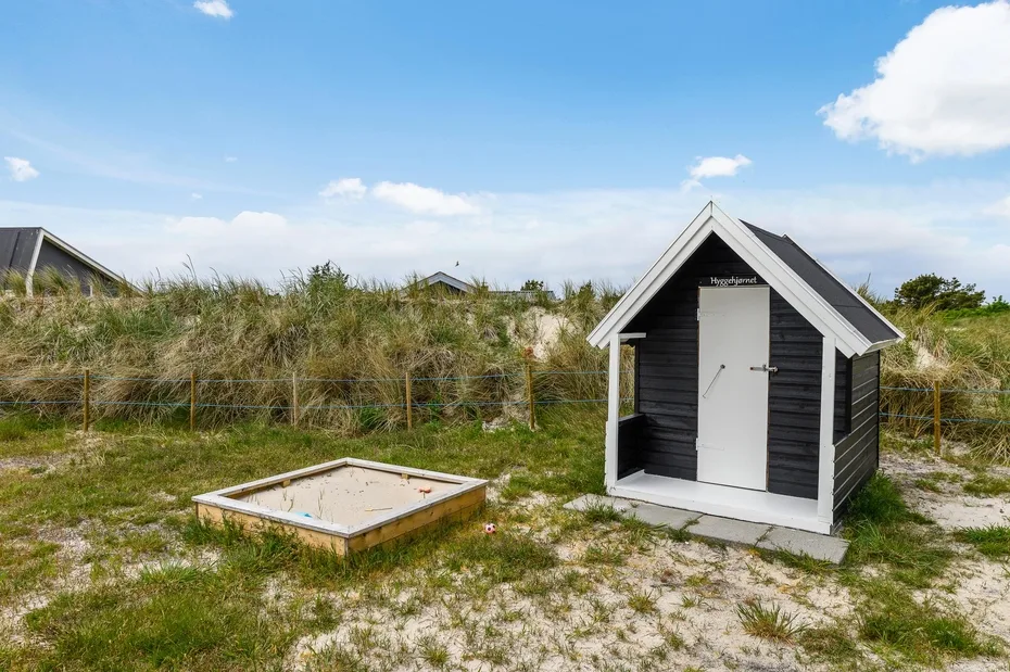 A small dark shed stands among grassy dunes beside a rectangular sand pit.