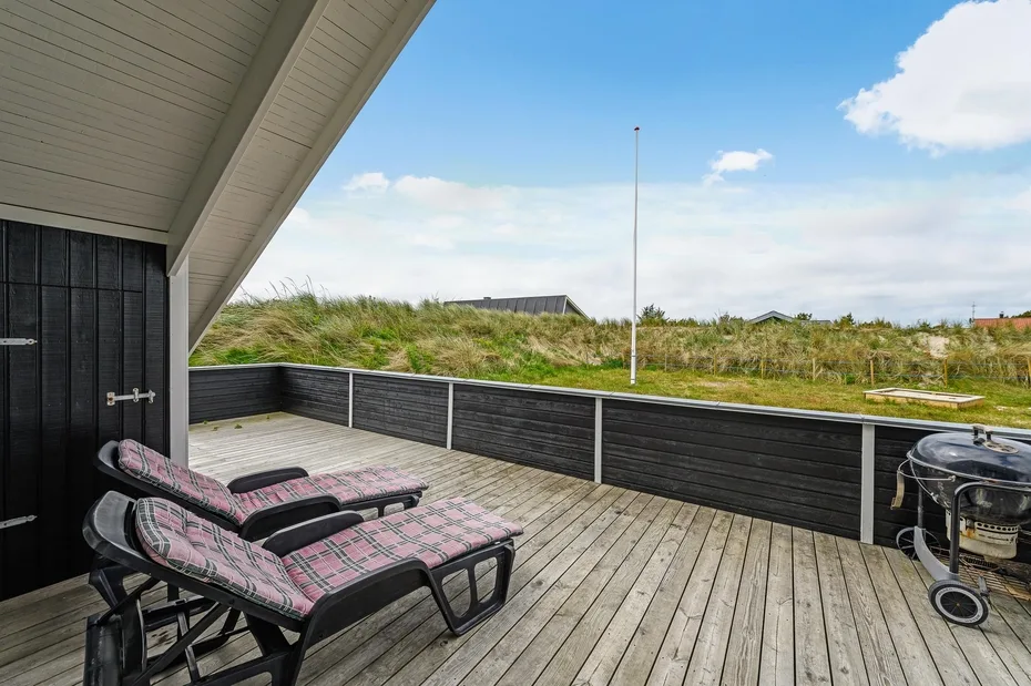 An outdoor deck with reclining seating overlooks grassy dunes beneath a blue sky.