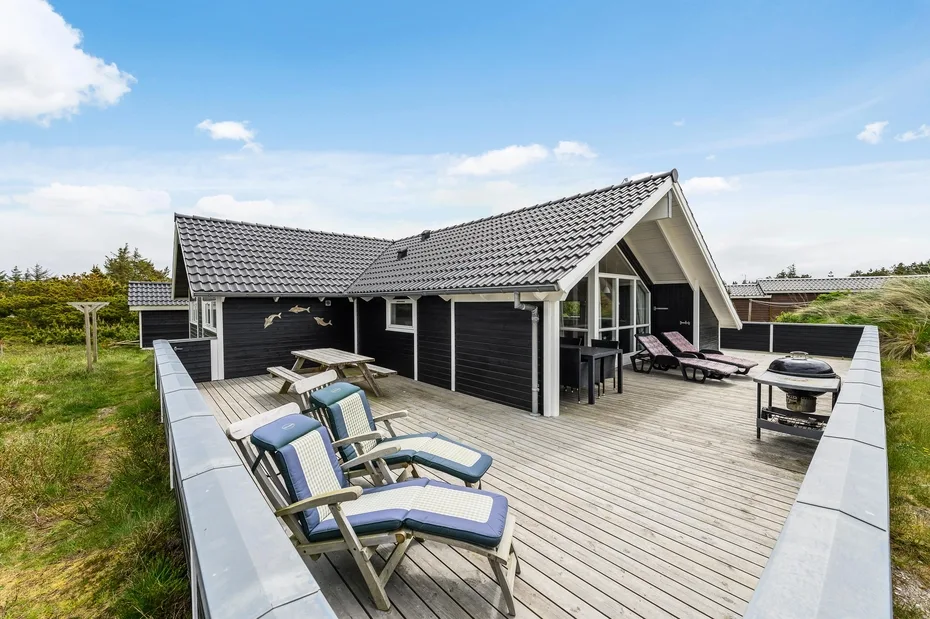 A spacious wooden deck with outdoor seating surrounds a dark-sided house under a blue sky.