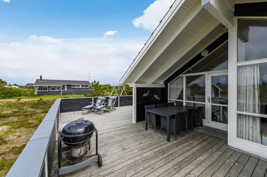 A wooden deck beside a house has outdoor seating, a dining table, and a barbecue grill.
