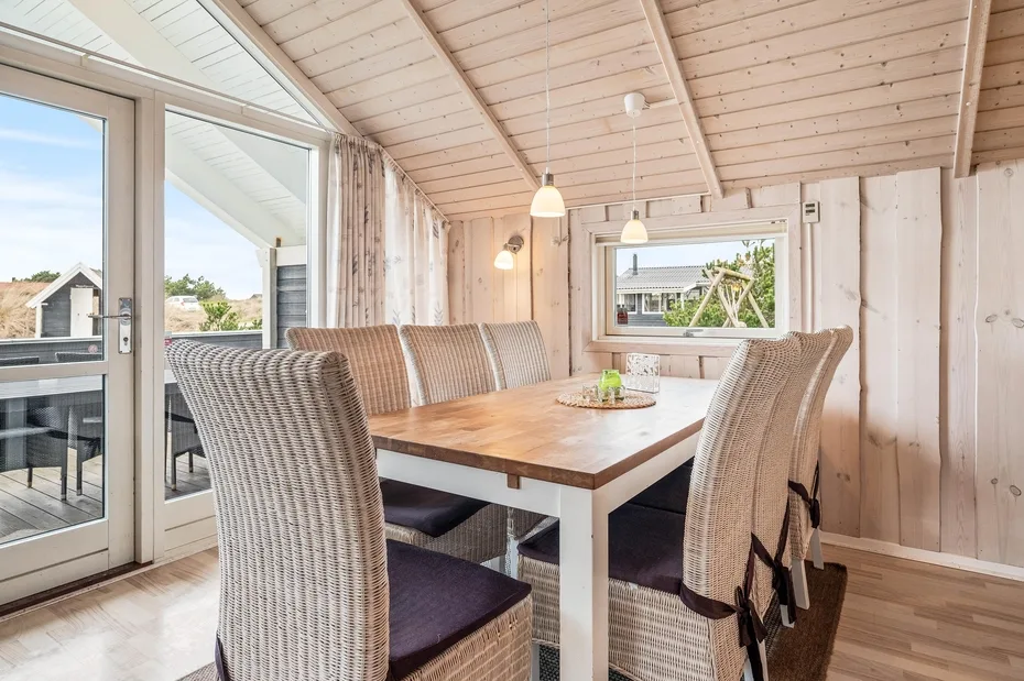 A dining area with a wooden table and woven seating beside large windows.