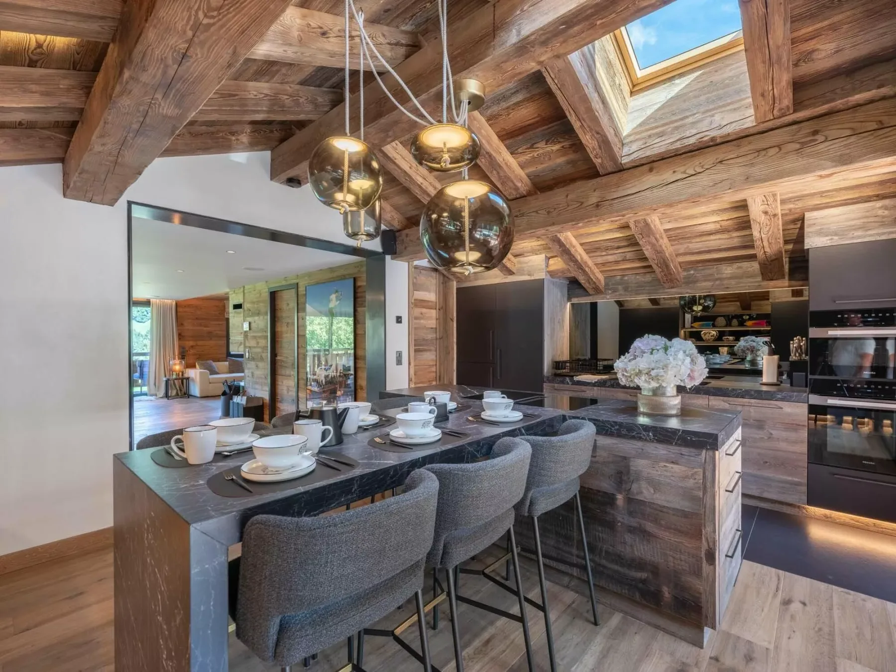 Una cocina y comedor de estilo rústico con una isla larga y luces colgantes bajo vigas de madera.