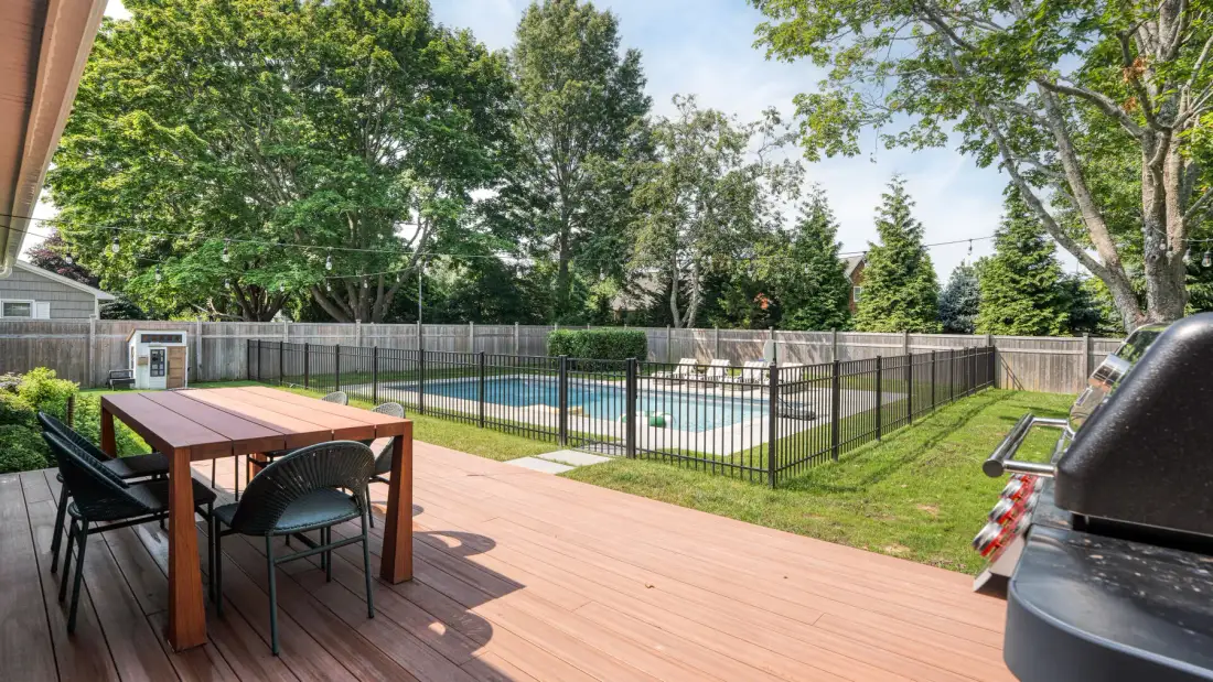 Una terraza de madera da a una piscina cercada, con asientos al aire libre y una parrilla cerca.