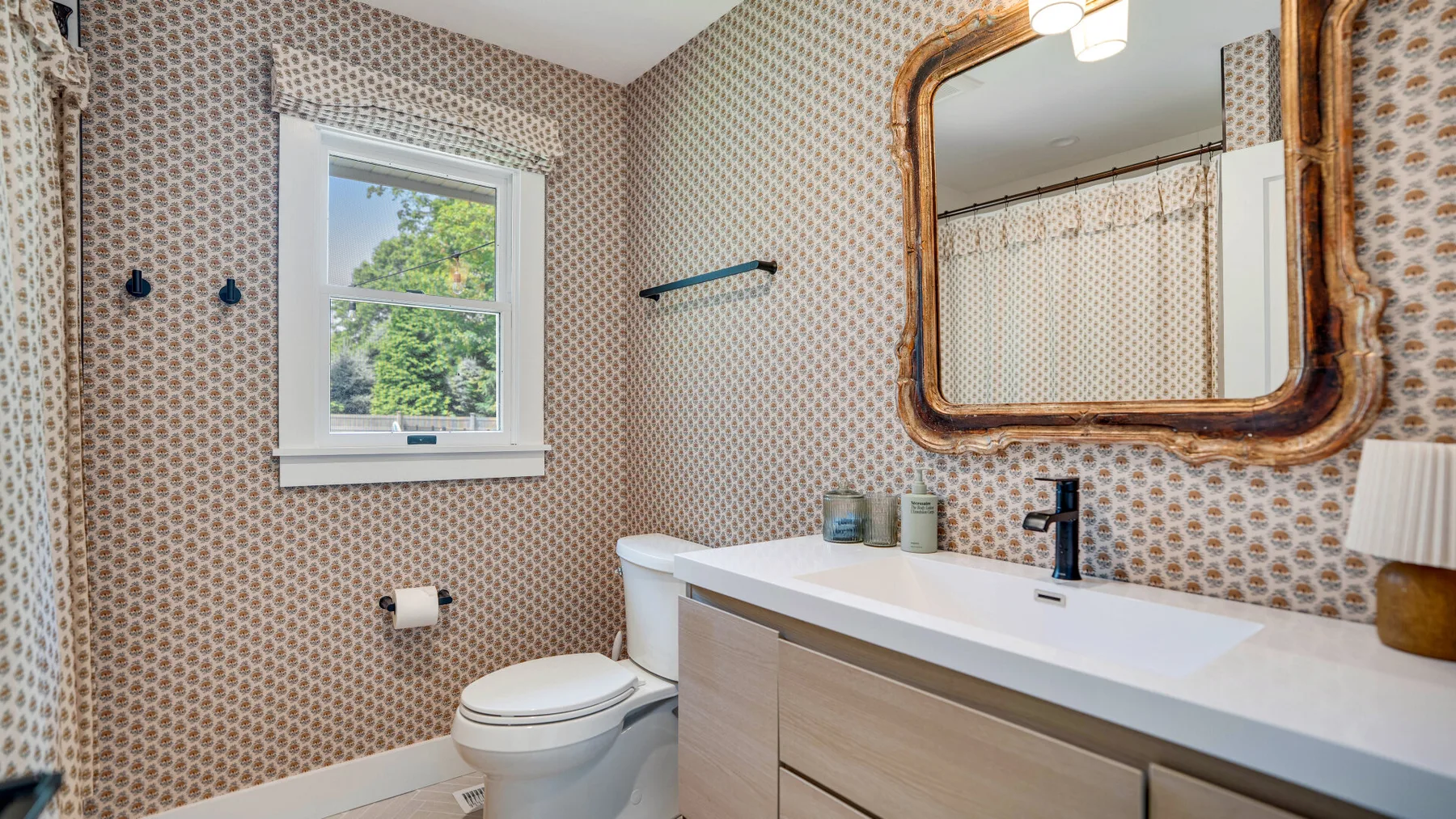 Un baño con paredes estampadas, tocador, espejo, inodoro y ventana.