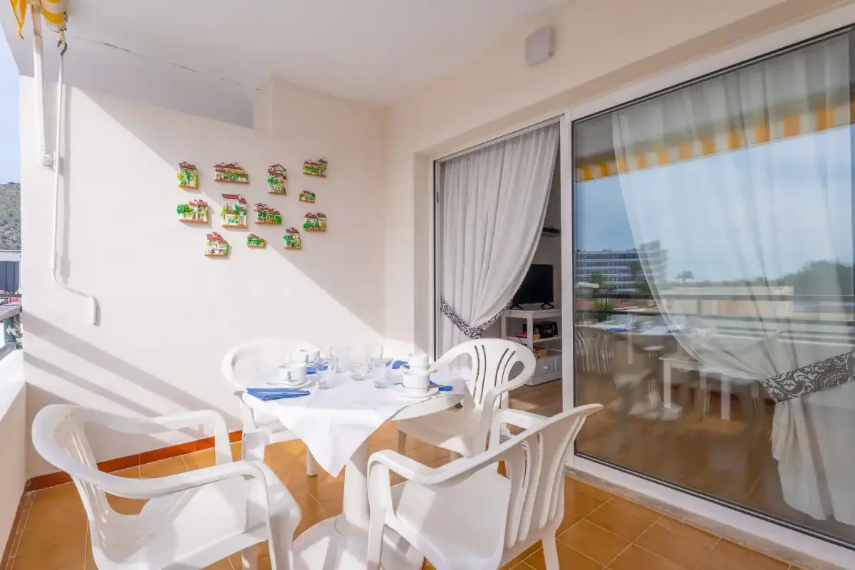 A sunlit balcony dining area has a table and white seating beside sliding glass doors.