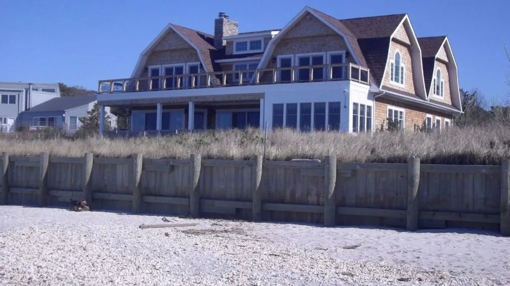 Una casa grande se alza detrás de una cerca de madera junto a una playa de arena.