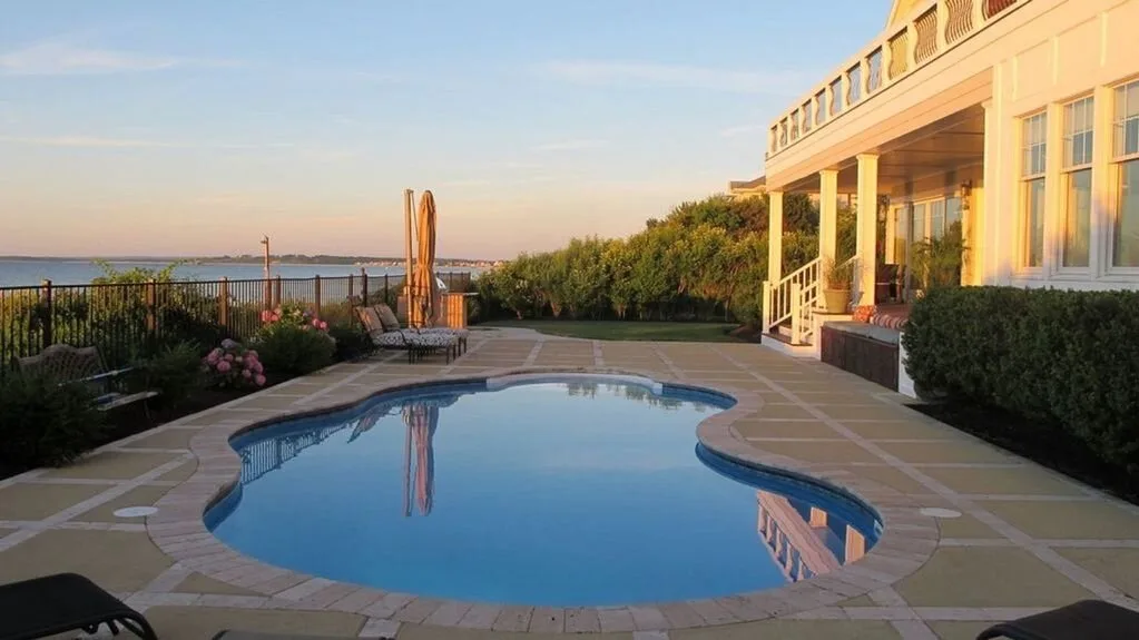 Outdoor pool beside a house, with patio paving and garden shrubs.