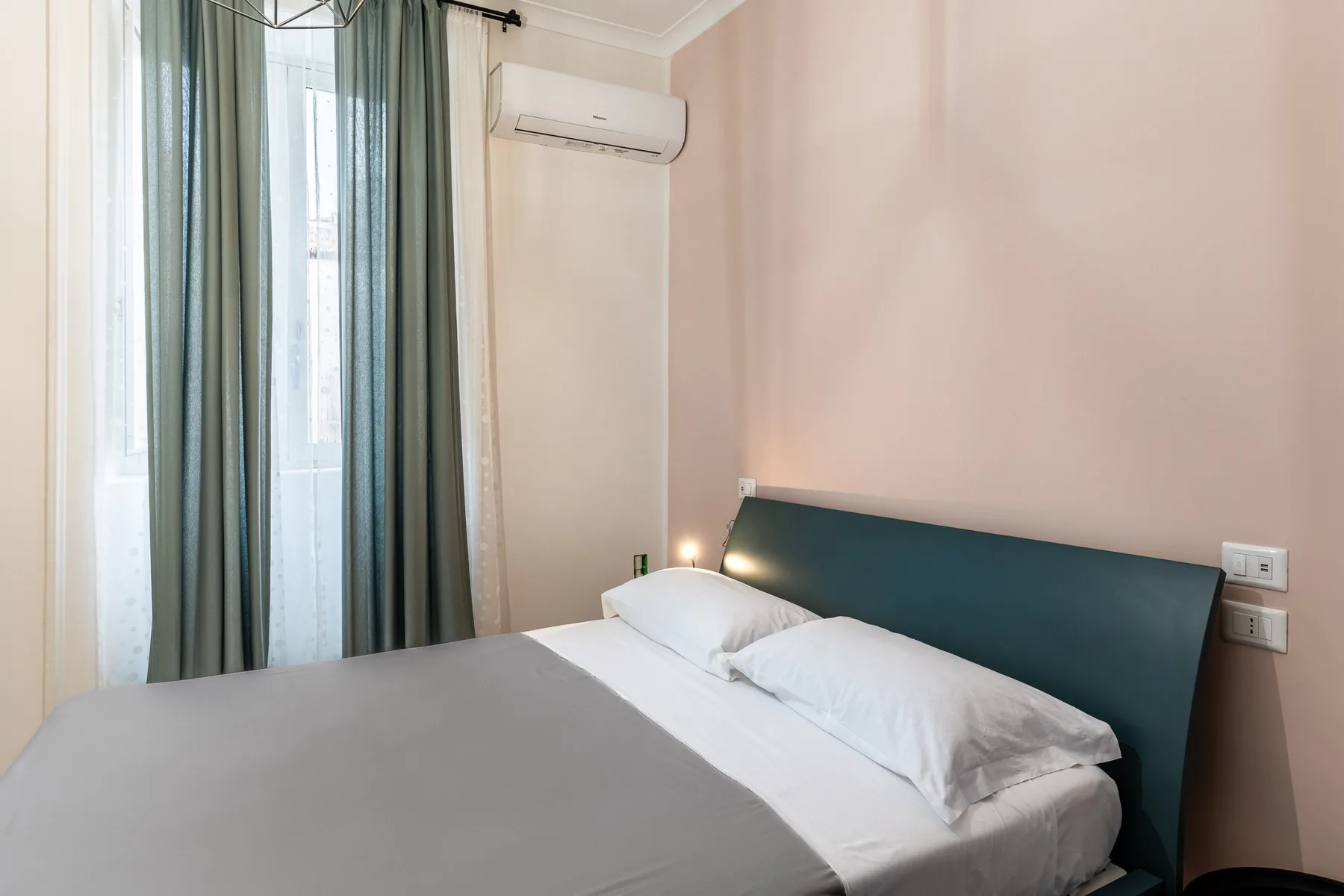 Un dormitorio con una cama, un cabecero verde azulado y ventanas altas con cortinas.