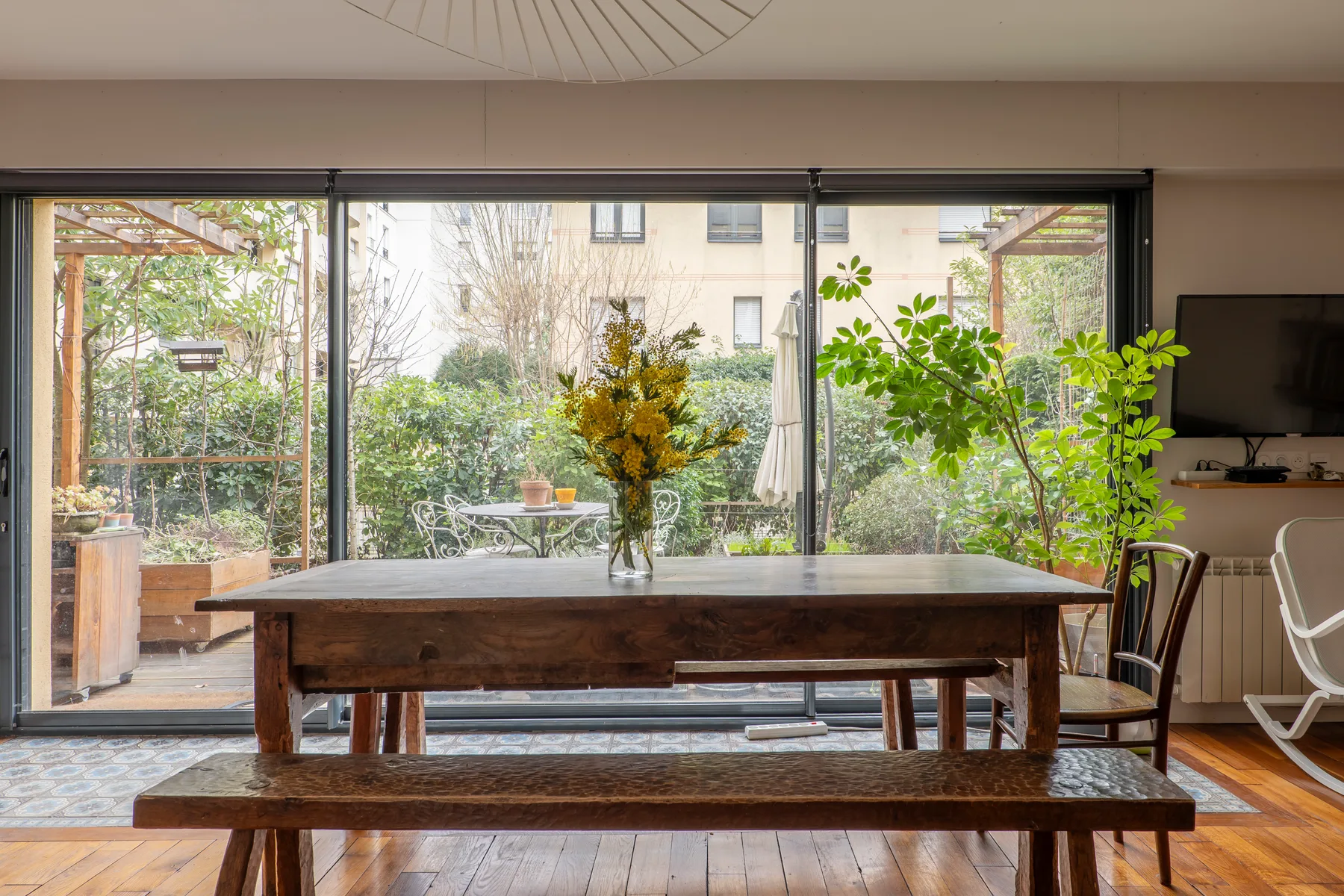 A wooden dining table faces large windows overlooking a leafy garden.