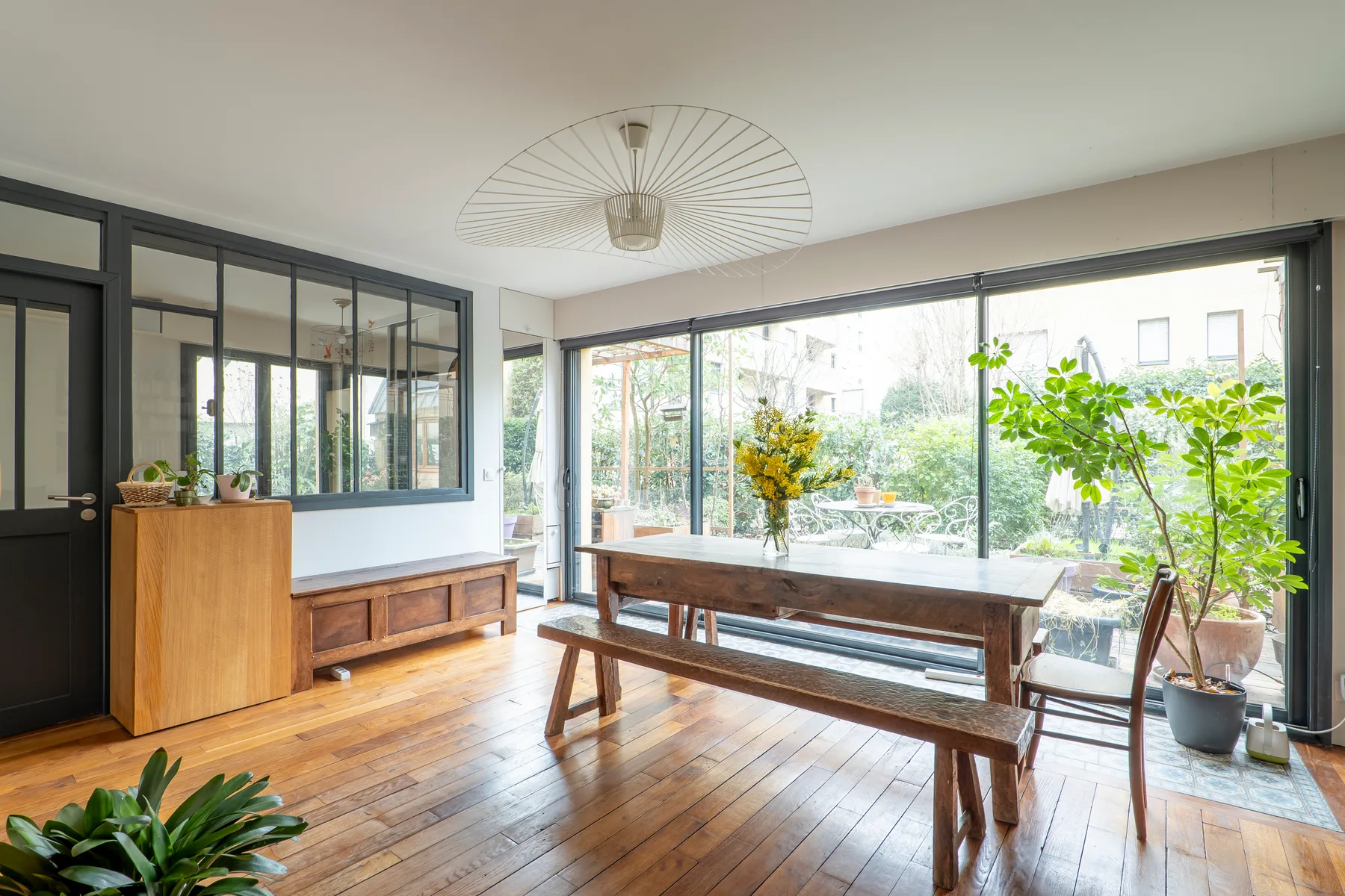 A bright dining area with a long wooden table beside large windows and greenery.