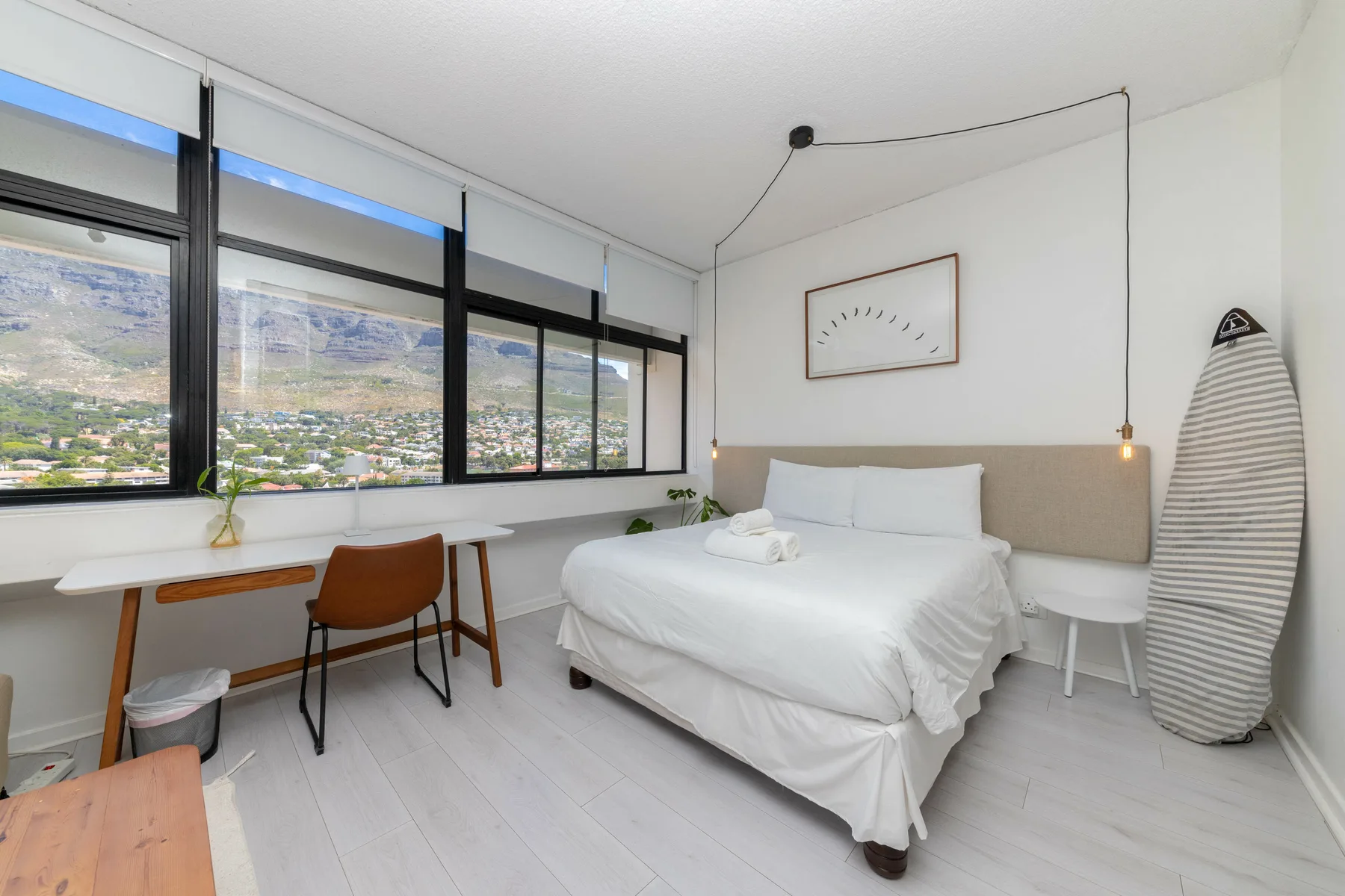 A bright bedroom with a bed, desk, and broad windows overlooking a hillside.