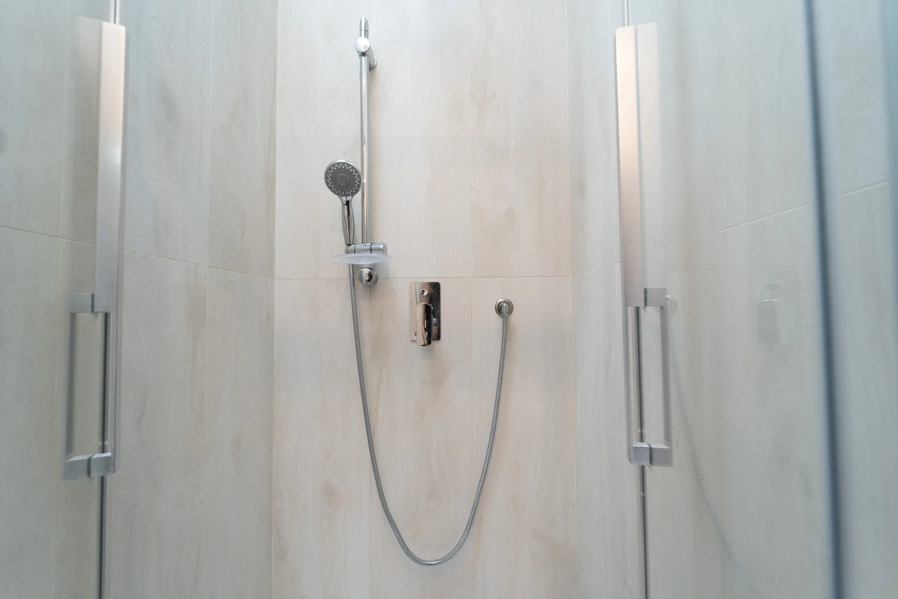 A tiled shower with a handheld showerhead and wall-mounted controls.