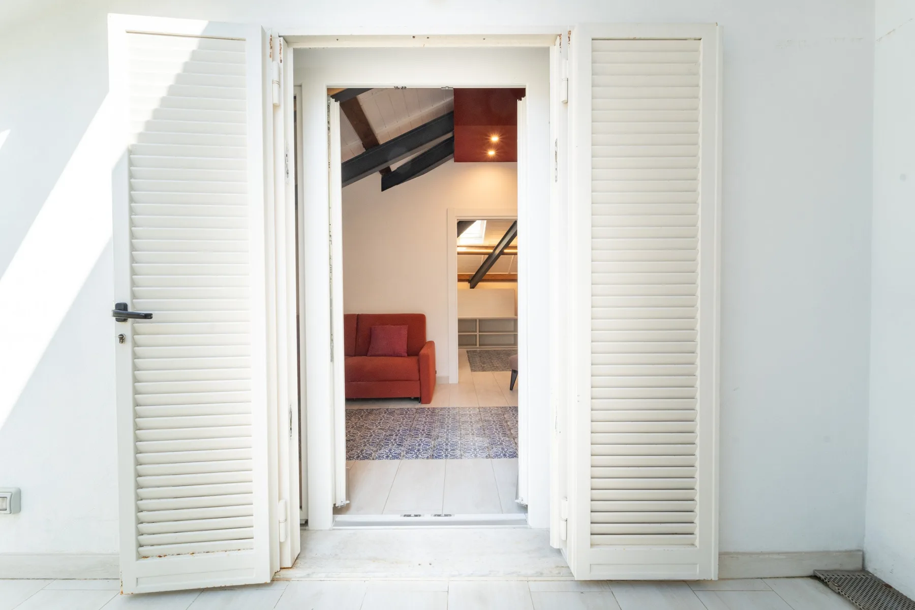 A bright room viewed through open white shutters, with a red seat and patterned rug.