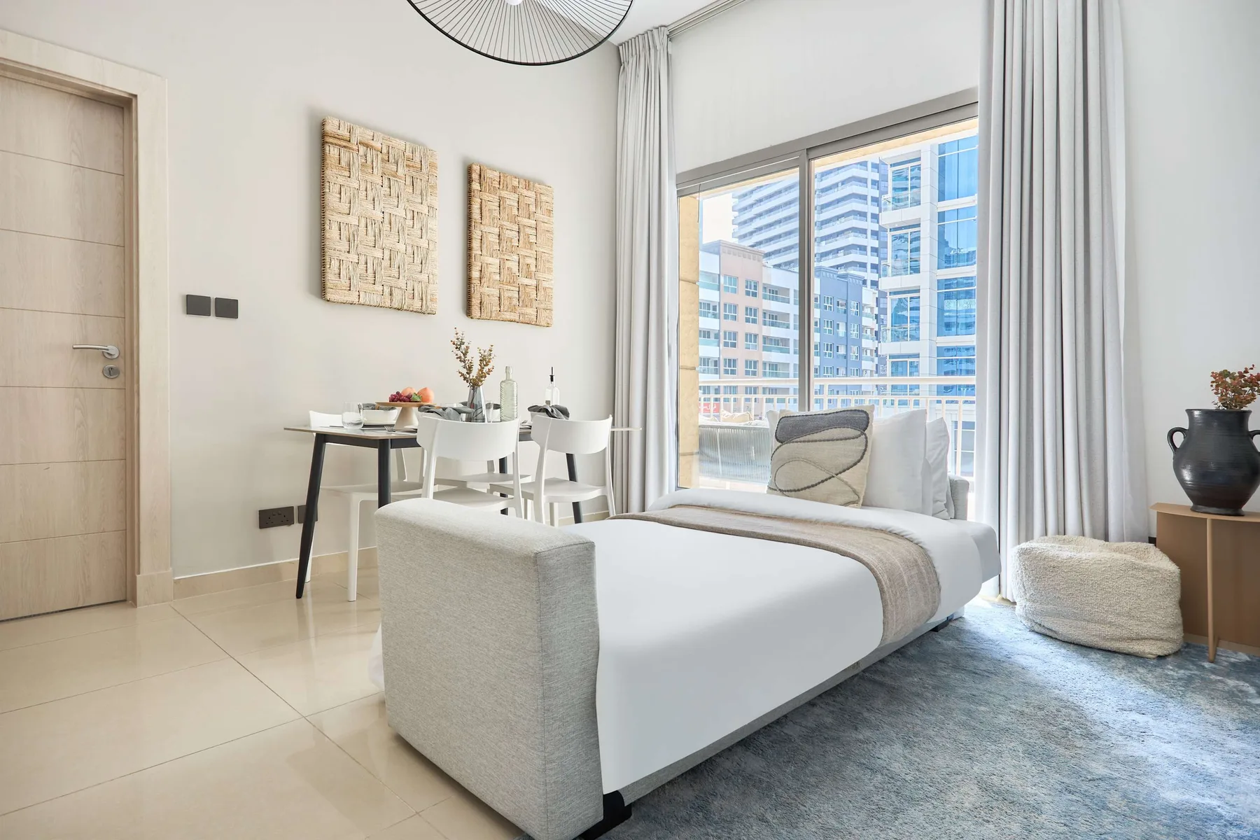 Un dormitorio luminoso con una cama, una mesa de comedor y grandes ventanas con edificios a la vista.