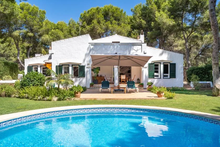 A white house with a shaded patio overlooks a swimming pool and green lawn.