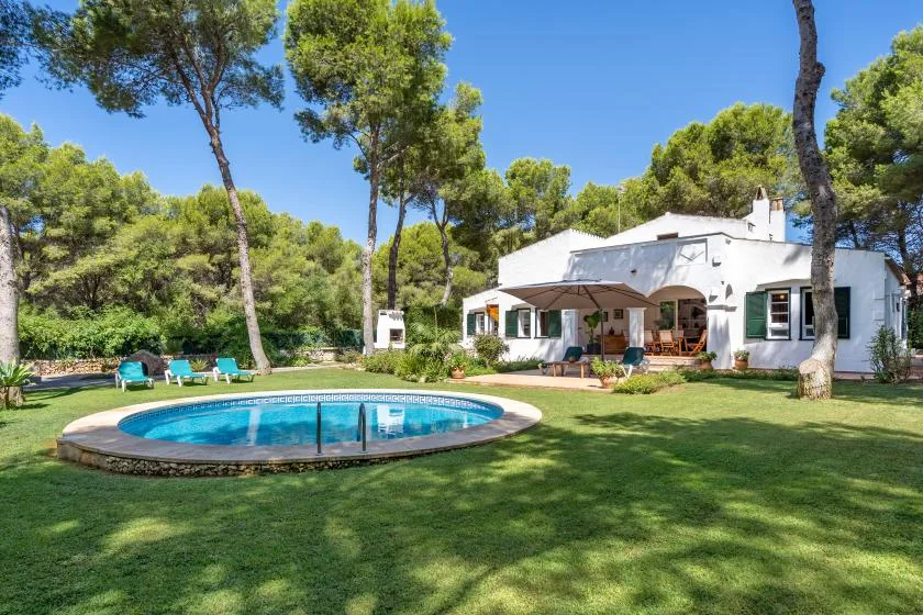 A white house and round swimming pool sit in a tree-lined garden.