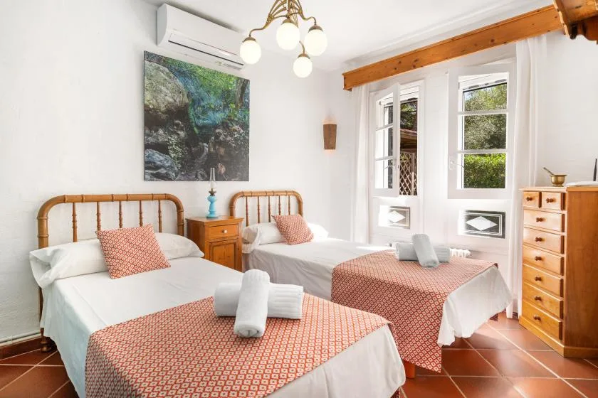 A bright bedroom with two beds, patterned covers, and windows overlooking greenery.