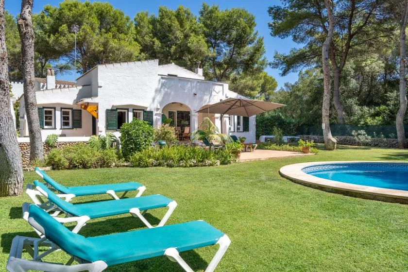 A white house opens onto a garden with sun loungers and a curved swimming pool.