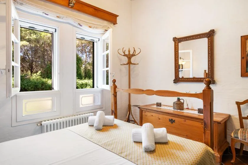 A bedroom with a wooden bed, folded towels, and open windows overlooking greenery.