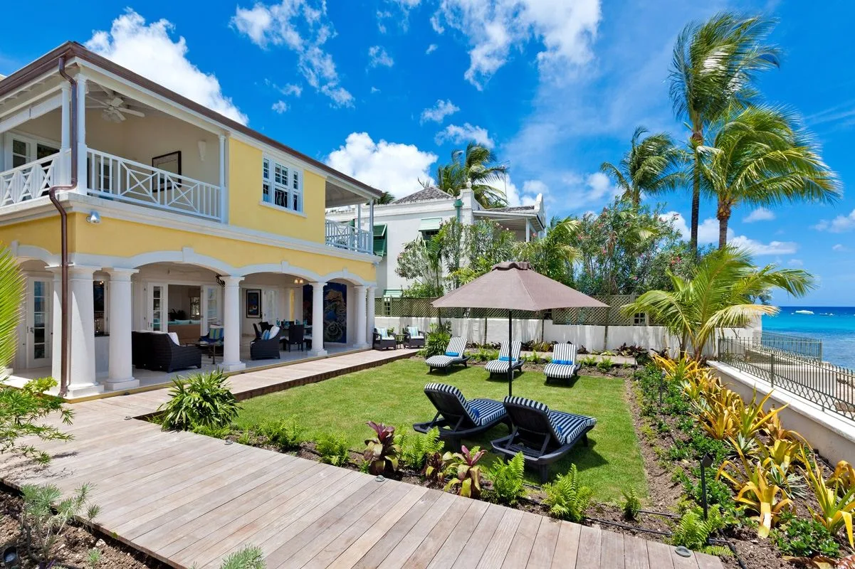 Una casa junto a la playa da a un césped con asientos al aire libre y palmeras.