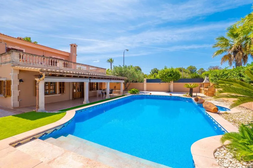 Una piscina junto a una casa, con un patio cubierto y palmeras.