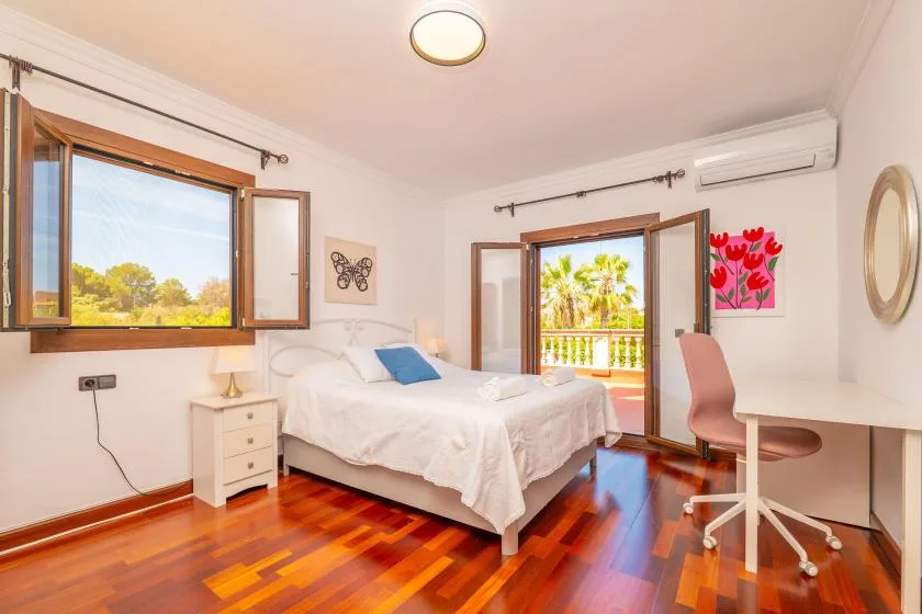 Un dormitorio luminoso con cama, escritorio y ventanas abiertas con vistas a la vegetación.