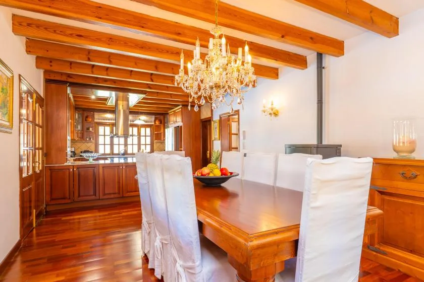 Una mesa de comedor de madera con asientos cubiertos está bajo vigas expuestas y una lámpara de araña.