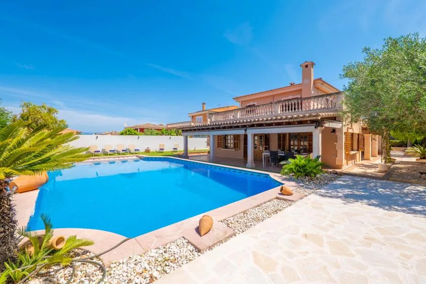 Piscina exterior junto a una casa, con un patio de piedra clara y plantas frondosas.