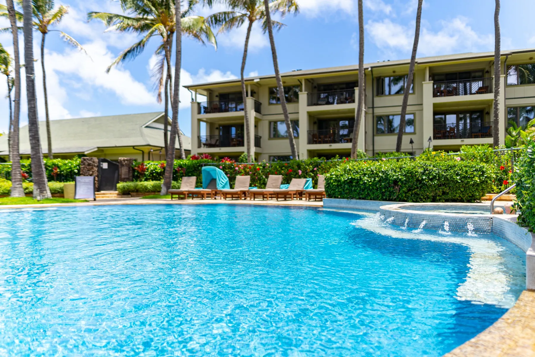 A swimming pool sits in front of a multi-story building and palm trees.