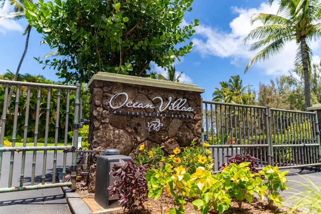 A stone entrance sign stands beside a metal fence among tropical plants and palm trees.