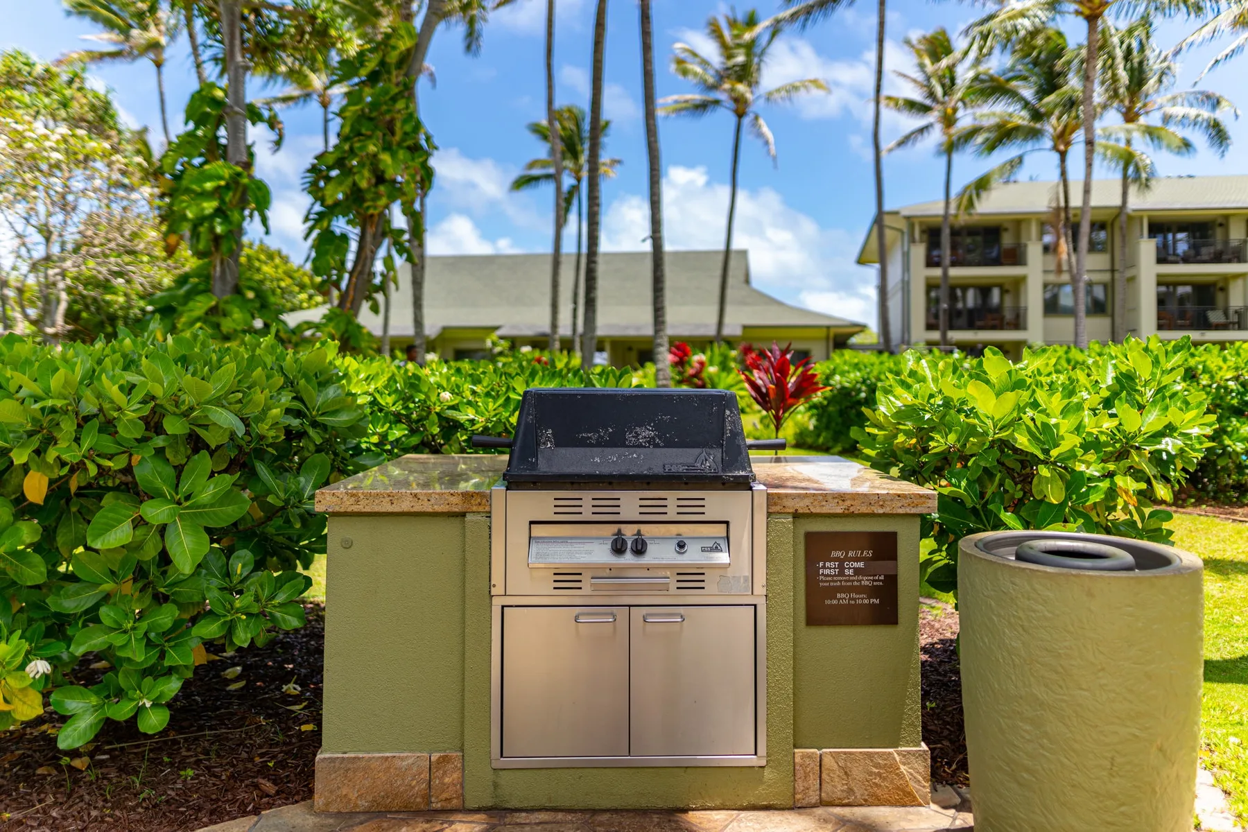 An outdoor grill stands among tropical landscaping near apartment buildings.