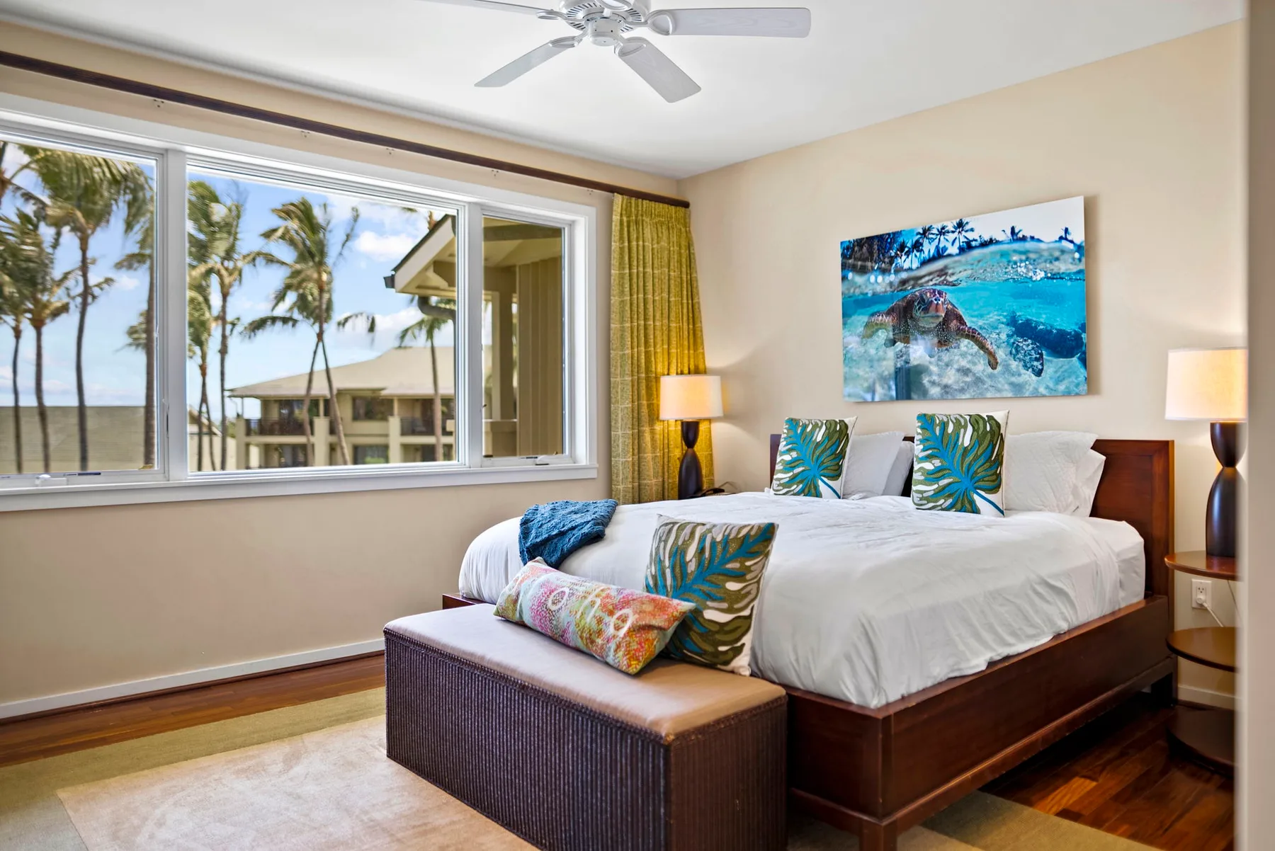 A bedroom with a large bed, colorful cushions, and a wide window overlooking palm trees.