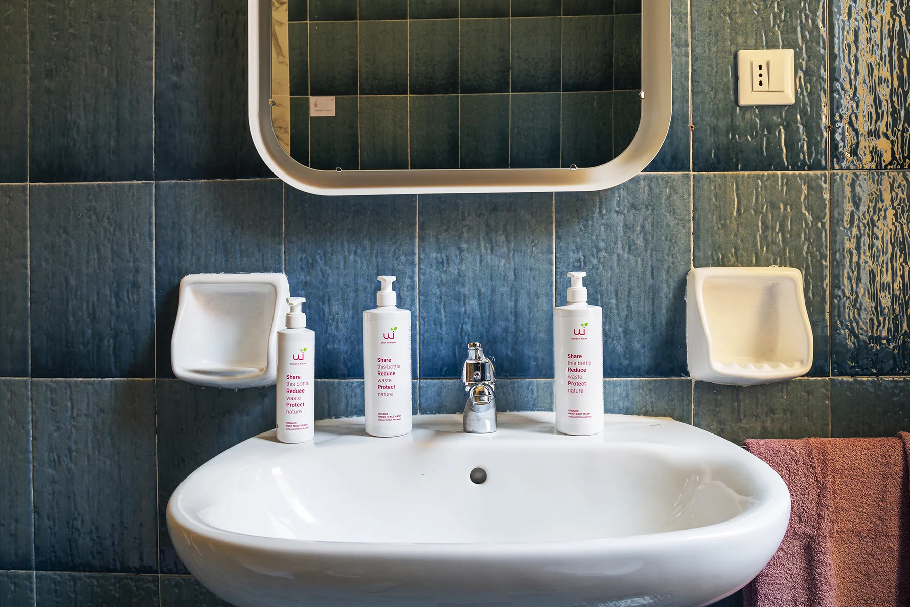 A bathroom sink sits beneath a mirror against dark tiled walls.