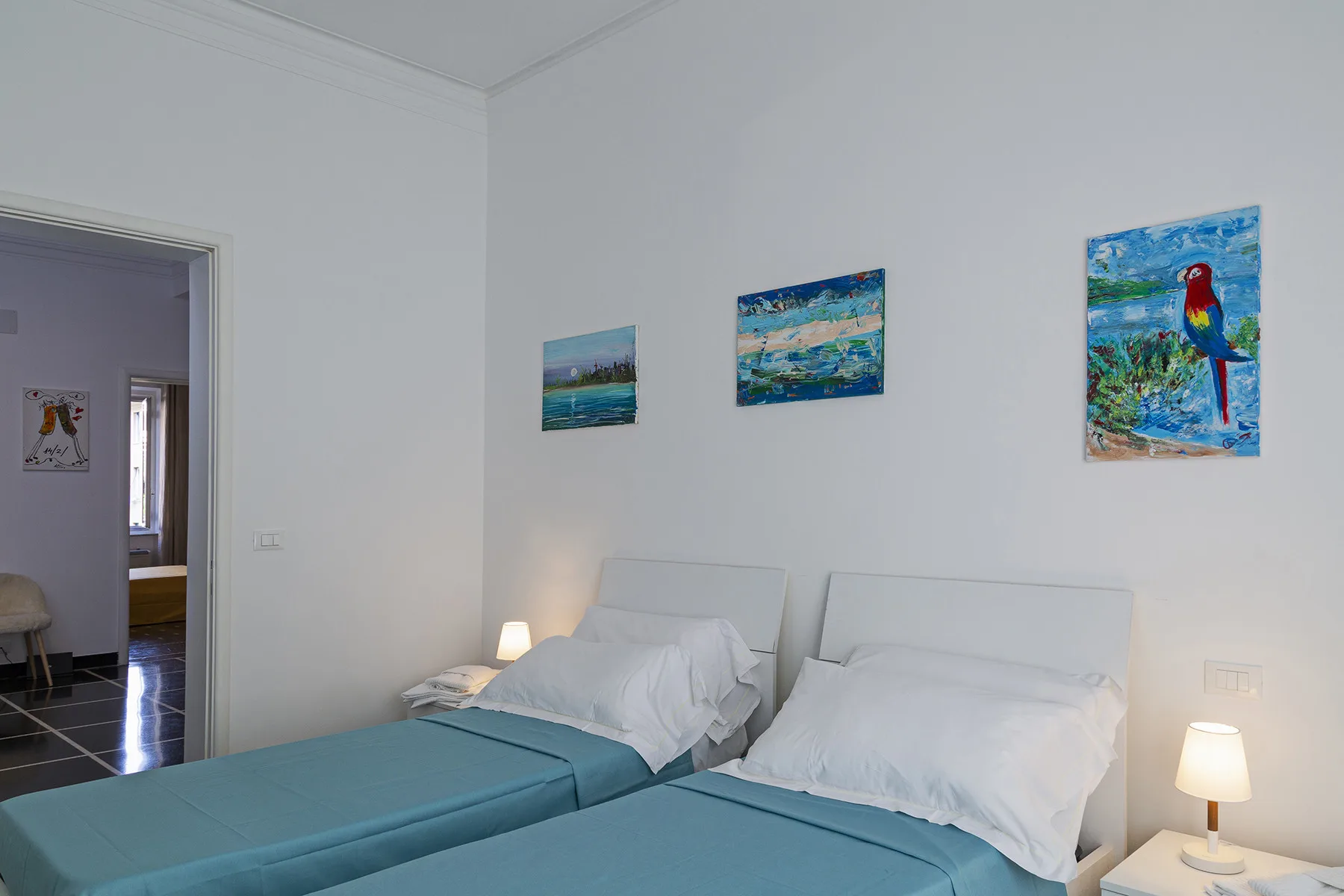 A bedroom with two beds, bedside lamps, and colorful paintings on the wall.